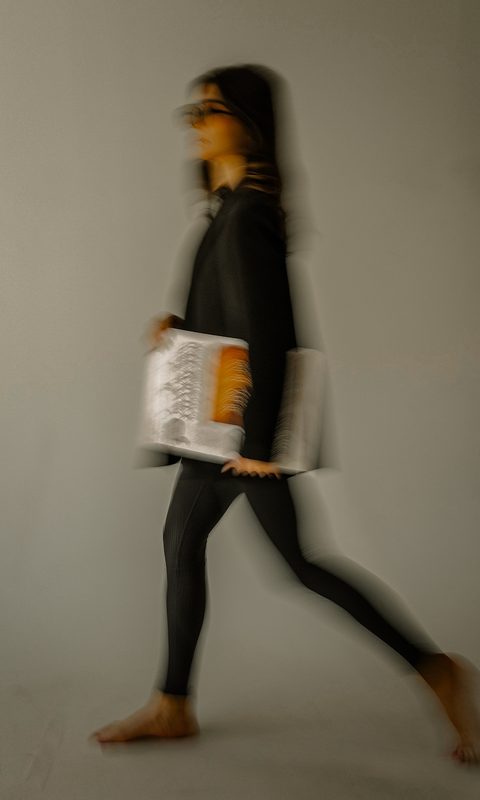 A motion-blurred figure walking across the studio with a mat under one arm
